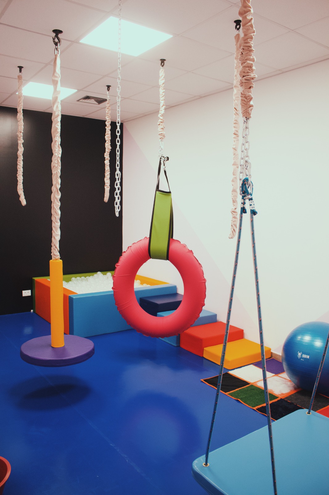 Sala de integração sensorial com balanços e argolas suspensas no teto, piscina de bolinhas colorida, pista de escalada e bola suíça azul, piso emborrachado azul vibrante, ambiente lúdico terapêutico, fotografia realista com iluminação natural
