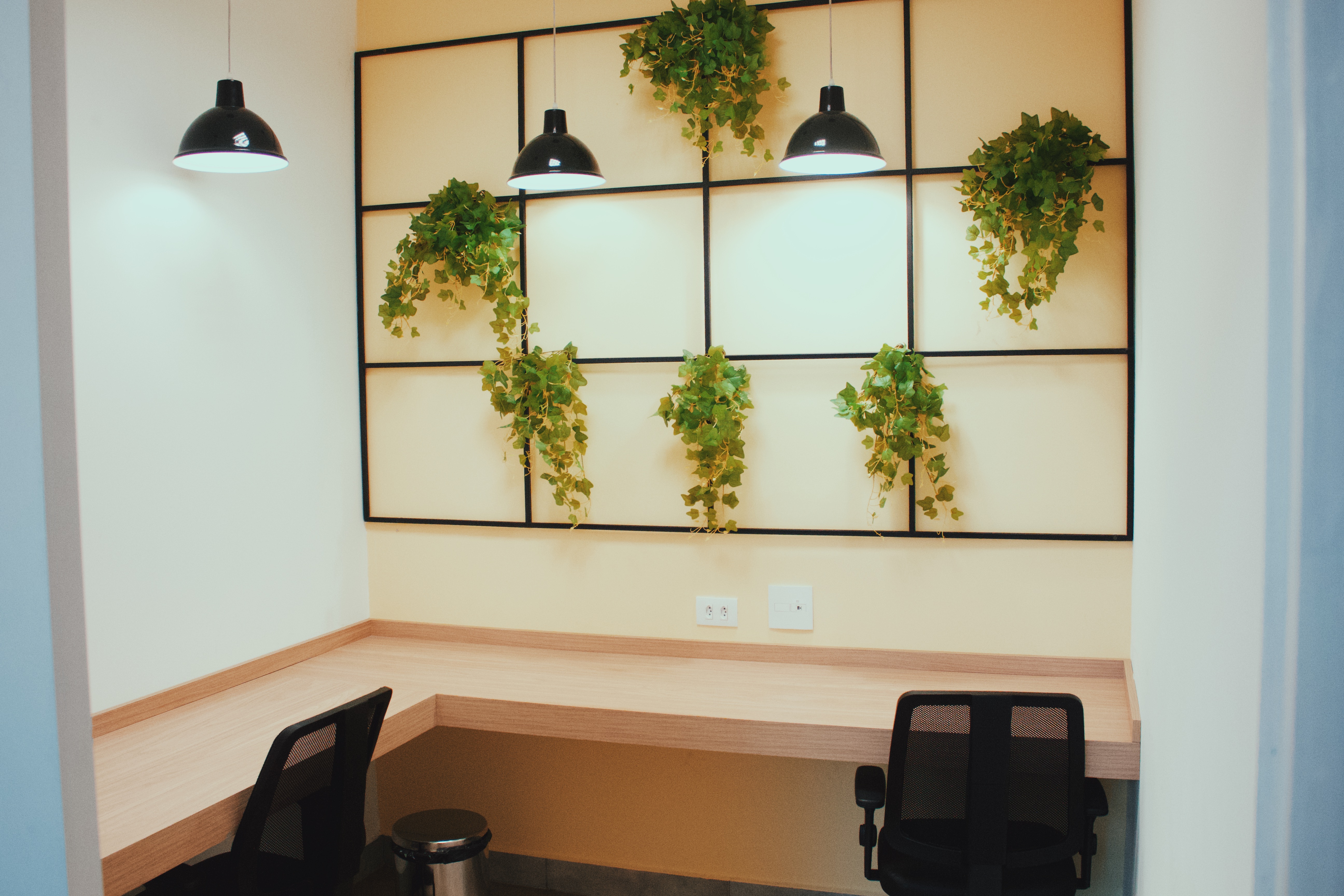 Espaço de coworking da clínica Abakids, mesas de madeira em bancada ao longo da parede, cadeiras de escritório pretas, luminárias pendentes e decoração com plantas suspensas, paleta bege e azul, fotografia realista de interior corporativo acolhedor