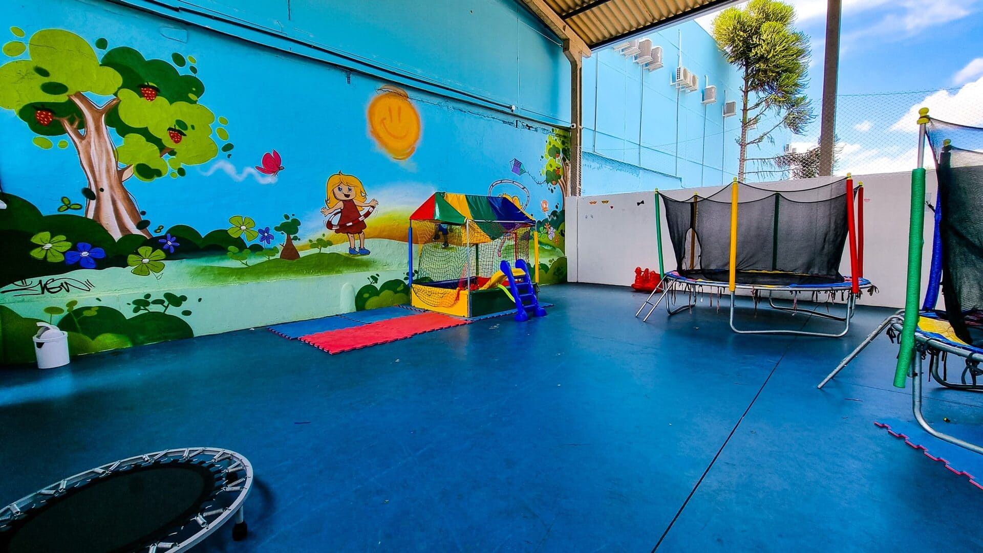 Parque coberto e lúdico da clínica com mural colorido pintado na parede, piscina de bolinhas, cama elástica e trampolins, piso azul emborrachado, fotografia realista de espaço infantil