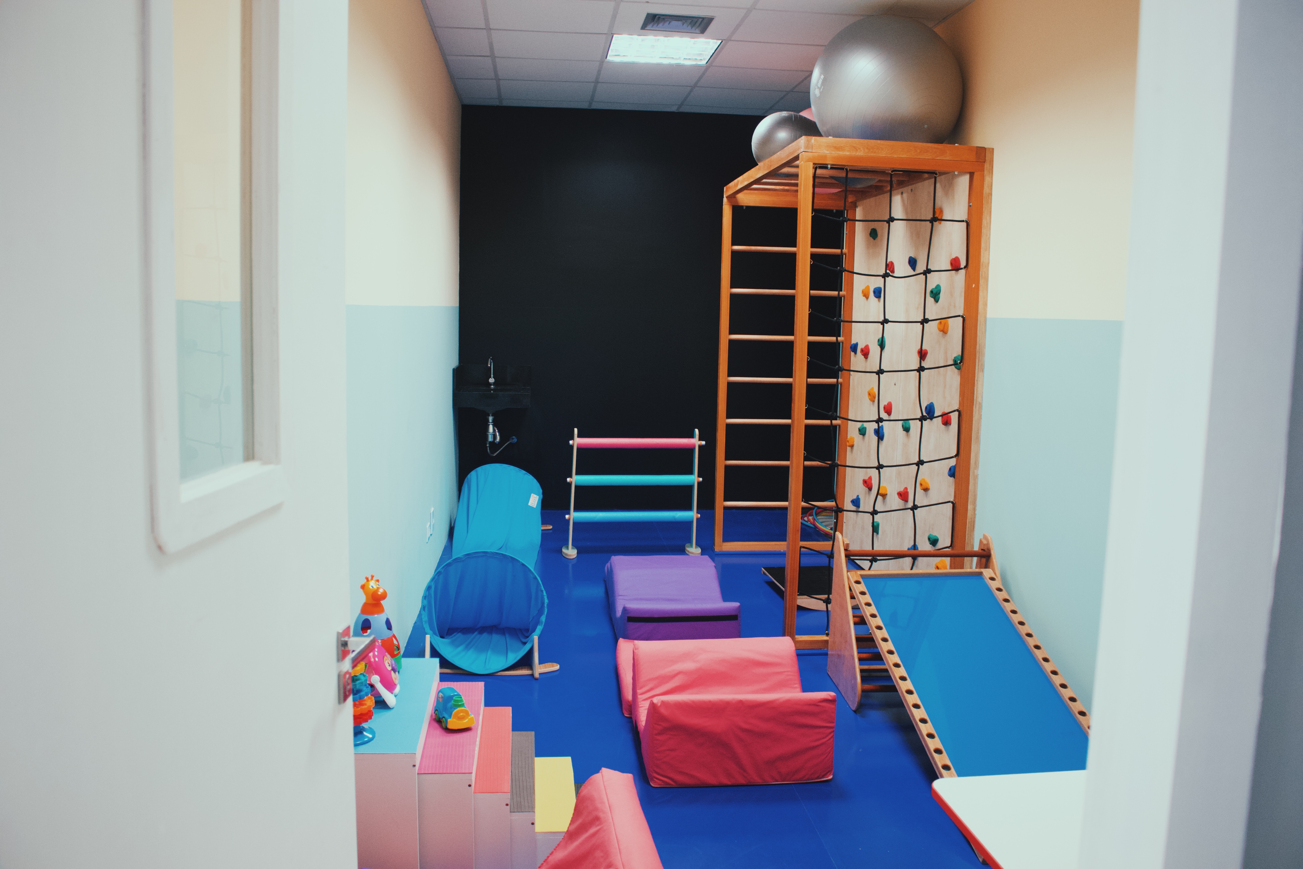 Sala de estimulação motora com parede de escalada colorida, escada sueca de madeira, bolas suíças e almofadas, piso emborrachado azul, fotografia realista de ambiente terapêutico