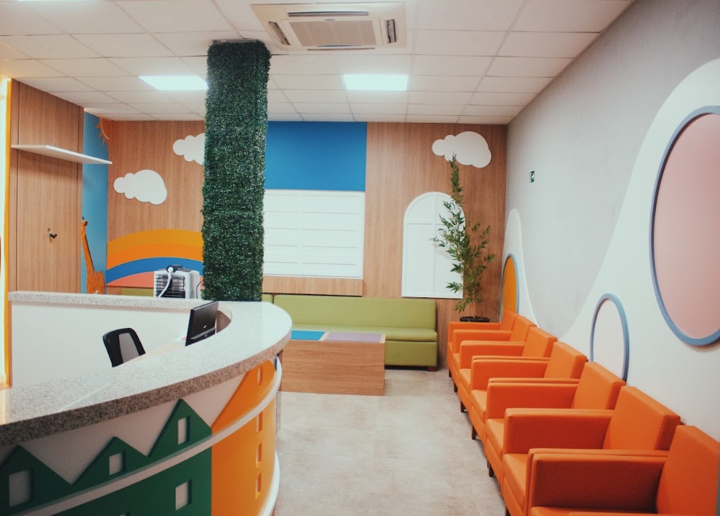 Recepção da clínica Abakids com balcão curvo decorado com ilustração de casinhas coloridas, parede com nuvens e arco-íris pintados, sofá verde e poltronas laranjas alinhadas, coluna revestida de folhagem artificial, piso claro e iluminação natural, fotografia realista de ambiente infantil acolhedor
