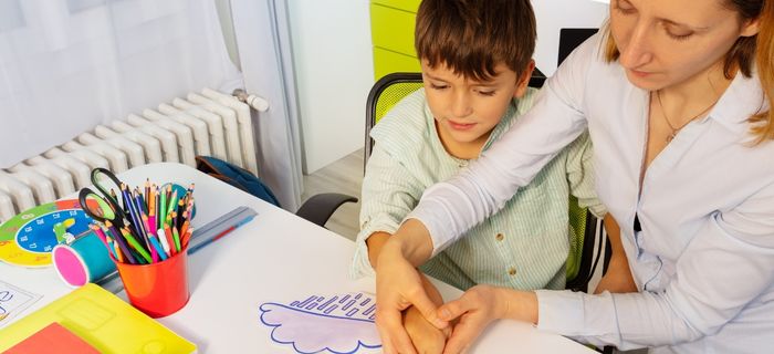Terapeuta auxiliando criança em atividade de terapia ABA, mãos guiando o desenho de uma nuvem com giz de cera sobre a mesa, materiais pedagógicos coloridos ao redor, ambiente clínico acolhedor, fotografia realista iluminação natural